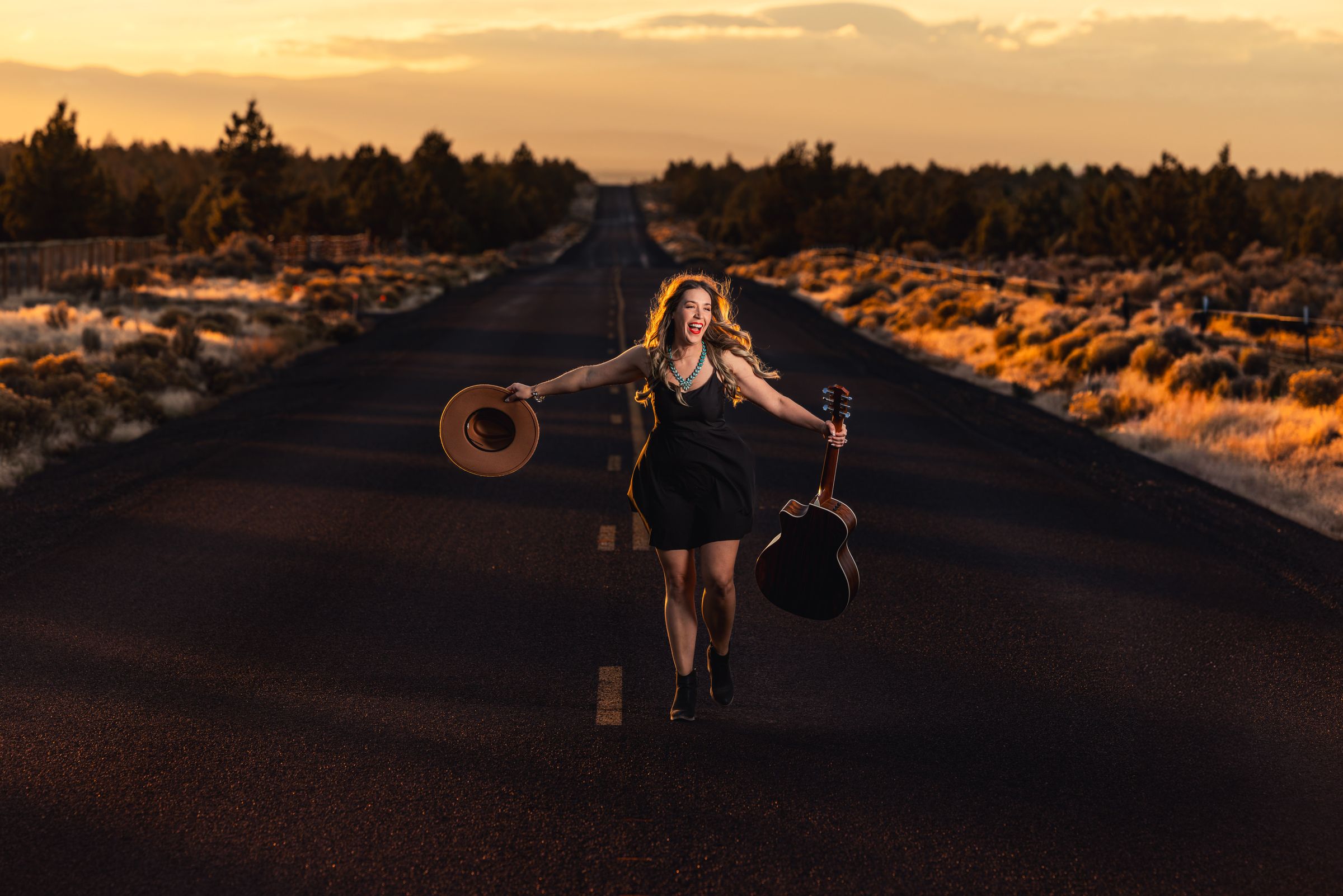 Aliki Samone raising her hat on an empty high-desert road at golden hour, guitar in hand
