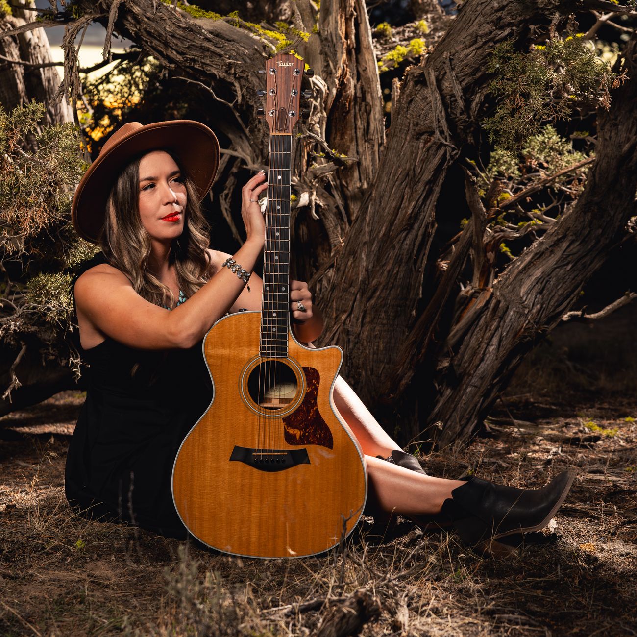 Aliki in a wide-brim hat, seated among juniper with her Taylor acoustic guitar