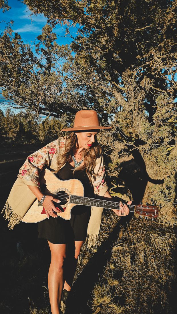 Aliki strumming her guitar in a hat and floral shawl