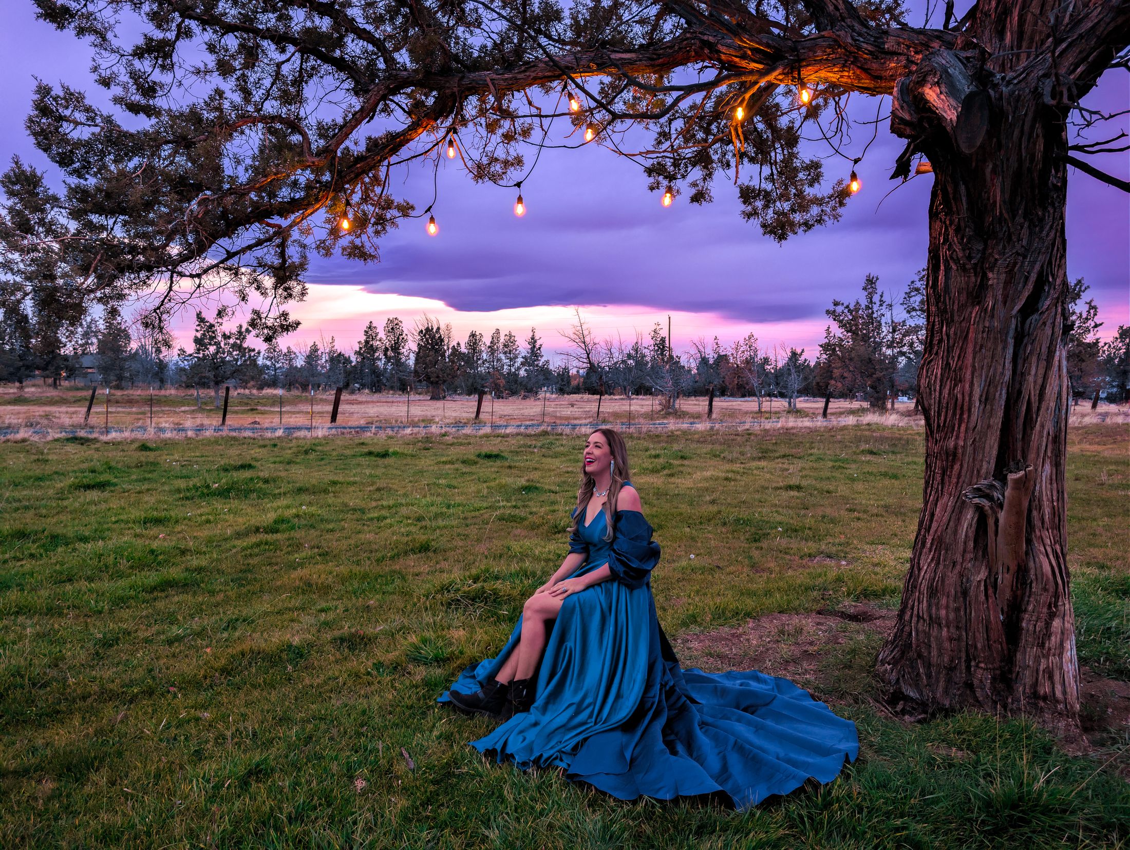 Aliki in a flowing teal gown beneath a lit juniper at a purple and pink sunset